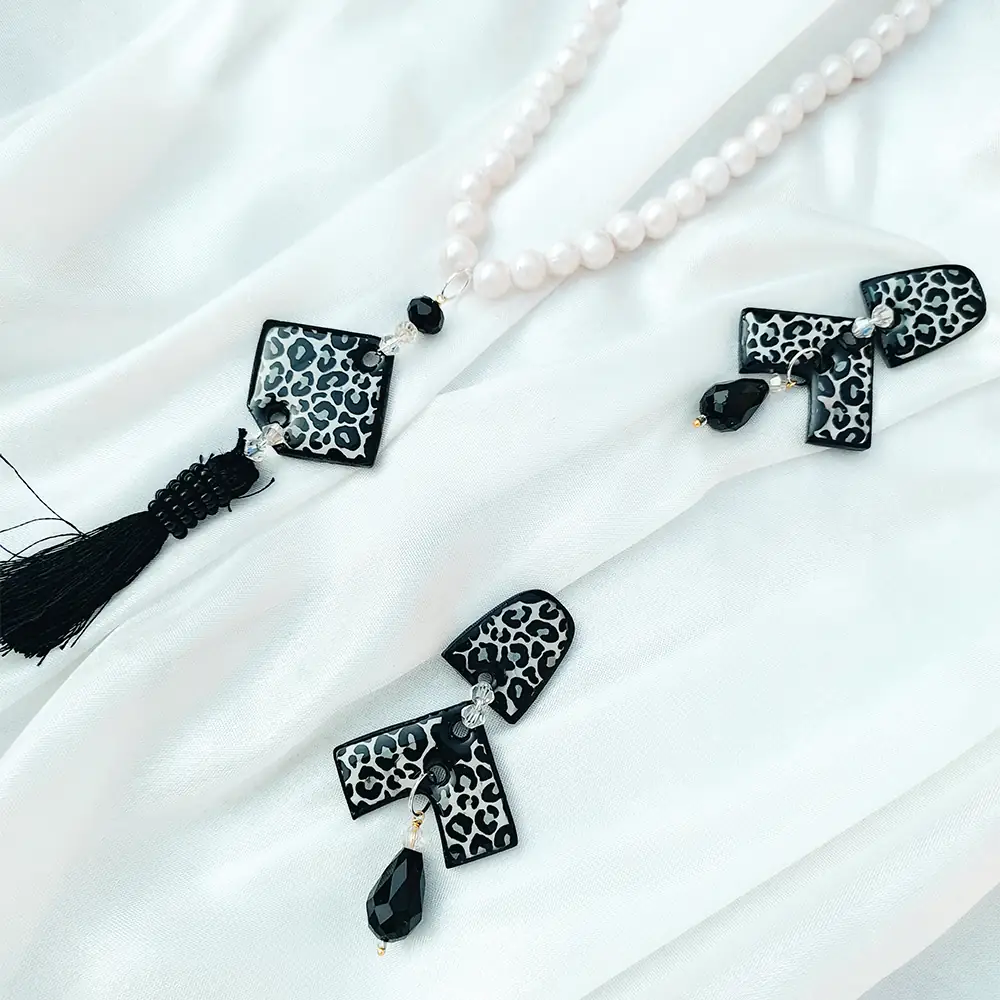 Black Leopard print resin pendant on a white pearl necklace next to matching geometric drop earrings on white silk fabric.