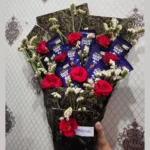 Close-up shot showing the arrangement of Dairy Milk chocolate bars surrounded by bright red roses and delicate white flowers.