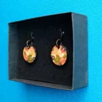 Pair of round clay mosaic floral earrings inside a black gift box against a blue wall.