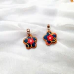 Pair of flower-shaped clay drop earrings with red blossoms and gold swirls resting on white silk.