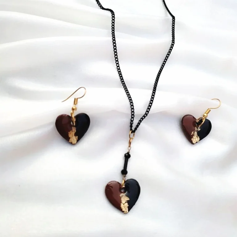A jewelry set consisting of split black and brown heart earrings and a matching pendant necklace on a black chain, resting on soft draped white silk fabric.