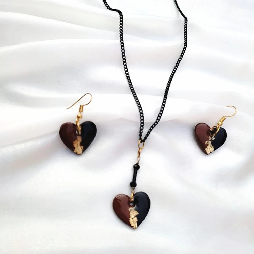 A jewelry set consisting of split black and brown heart earrings and a matching pendant necklace on a black chain, resting on soft draped white silk fabric.