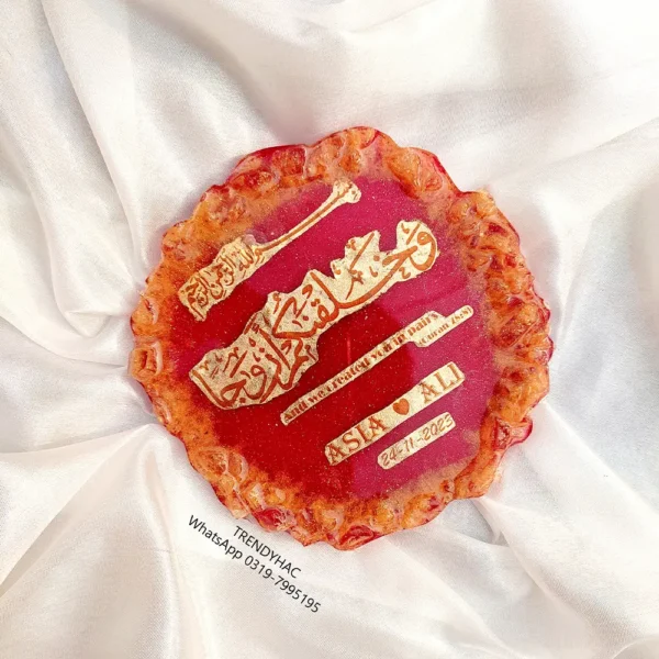 Flat lay view of a circular red resin wedding plaque with gold calligraphy and the names Asia and Ali on white satin fabric.