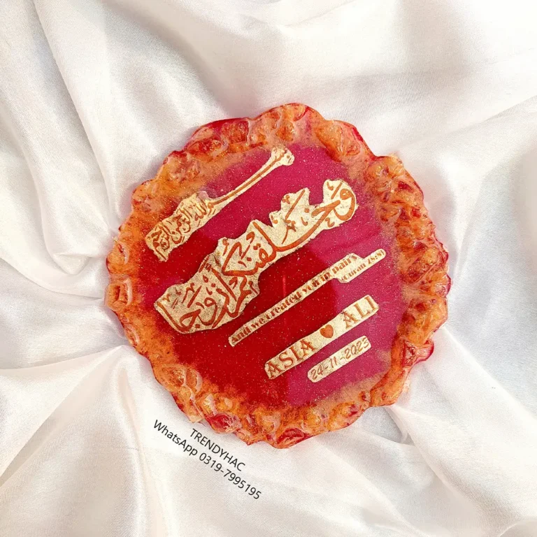 Flat lay view of a circular red resin wedding plaque with gold calligraphy and the names Asia and Ali on white satin fabric.