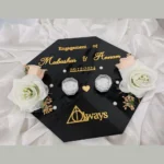 Flat lay view of a personalized black acrylic engagement ring tray with gold lettering, clear crystal ring boxes, and white roses.