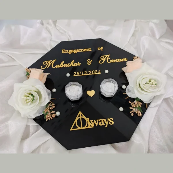Flat lay view of a personalized black acrylic engagement ring tray with gold lettering, clear crystal ring boxes, and white roses.