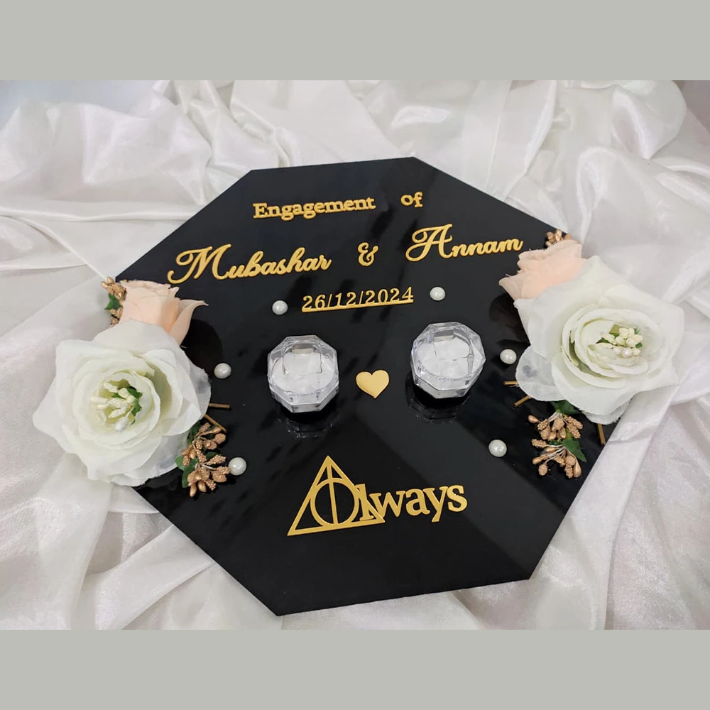 Flat lay view of a personalized black acrylic engagement ring tray with gold lettering, clear crystal ring boxes, and white roses.