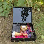 Open black chocolate gift box with assorted jewellery, perfume, letter, flower, chocolates and heart-shaped chocolate pack