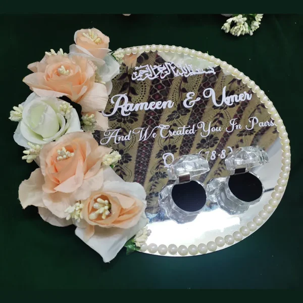 Overhead view of a round mirror acrylic Nikah ring tray featuring white Islamic calligraphy, personalized names, a pearl border, and peach roses.