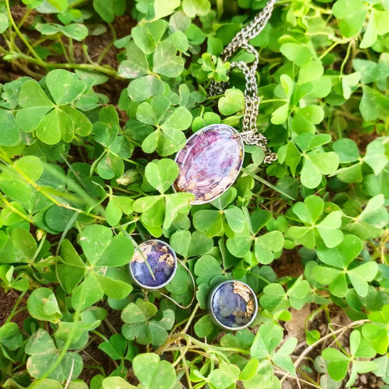 Front view of a Preserved Dried Flower jewelry set with an oval pendant necklace and round stud earrings containing purple petals and gold flakes on black velvet.