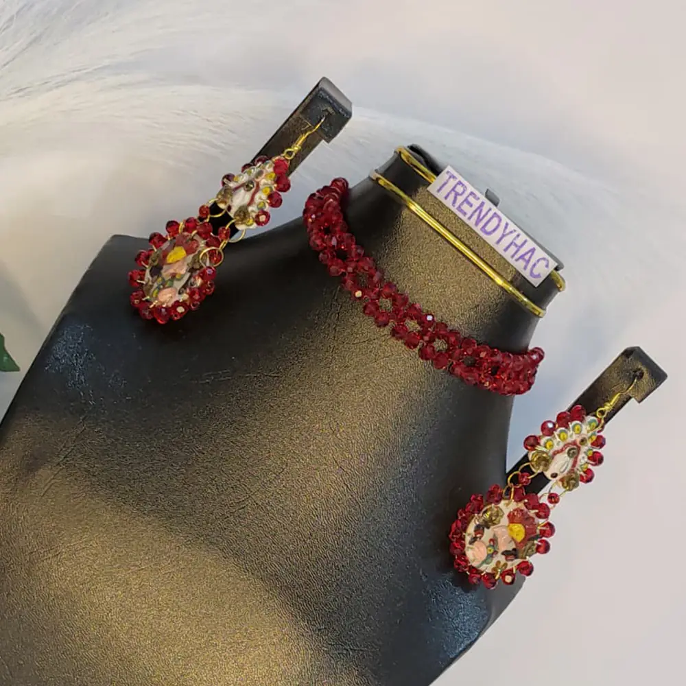 A deep red woven crystal beaded choker necklace displayed on a black bust alongside a matching tiered clay drop earring with hand-painted yellow and red floral details.