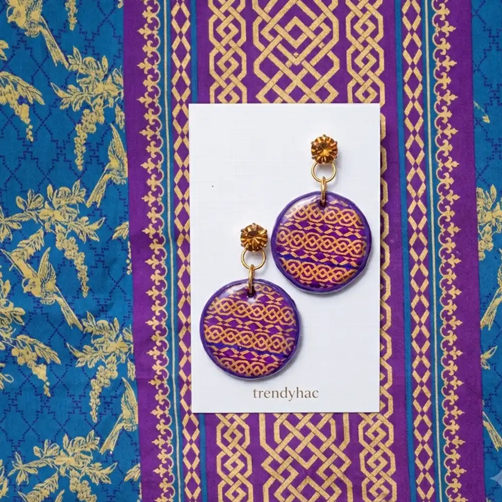Round purple clay drop earrings with intricate gold geometric interlocking line patterns on a white trendyhac card over matching purple and gold fabric.