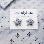Flat lay view of light grey star-shaped clay stud earrings with hand-painted black starburst patterns on a white TRENDYHAC display card matching the fabric background.