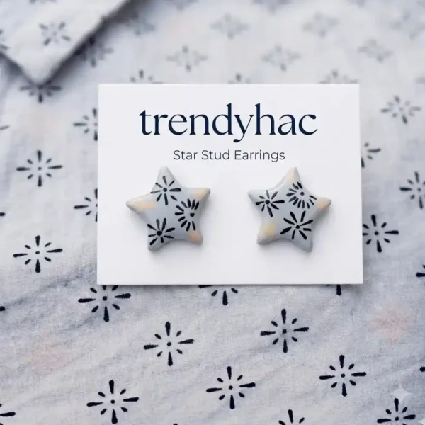 Flat lay view of light grey star-shaped clay stud earrings with hand-painted black starburst patterns on a white TRENDYHAC display card matching the fabric background.
