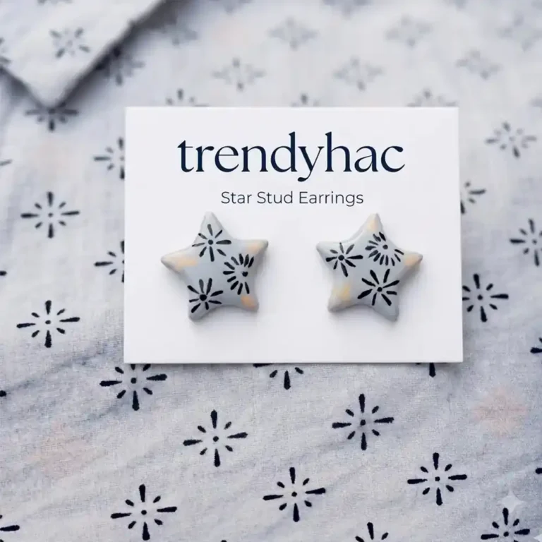 Flat lay view of light grey star-shaped clay stud earrings with hand-painted black starburst patterns on a white TRENDYHAC display card matching the fabric background.
