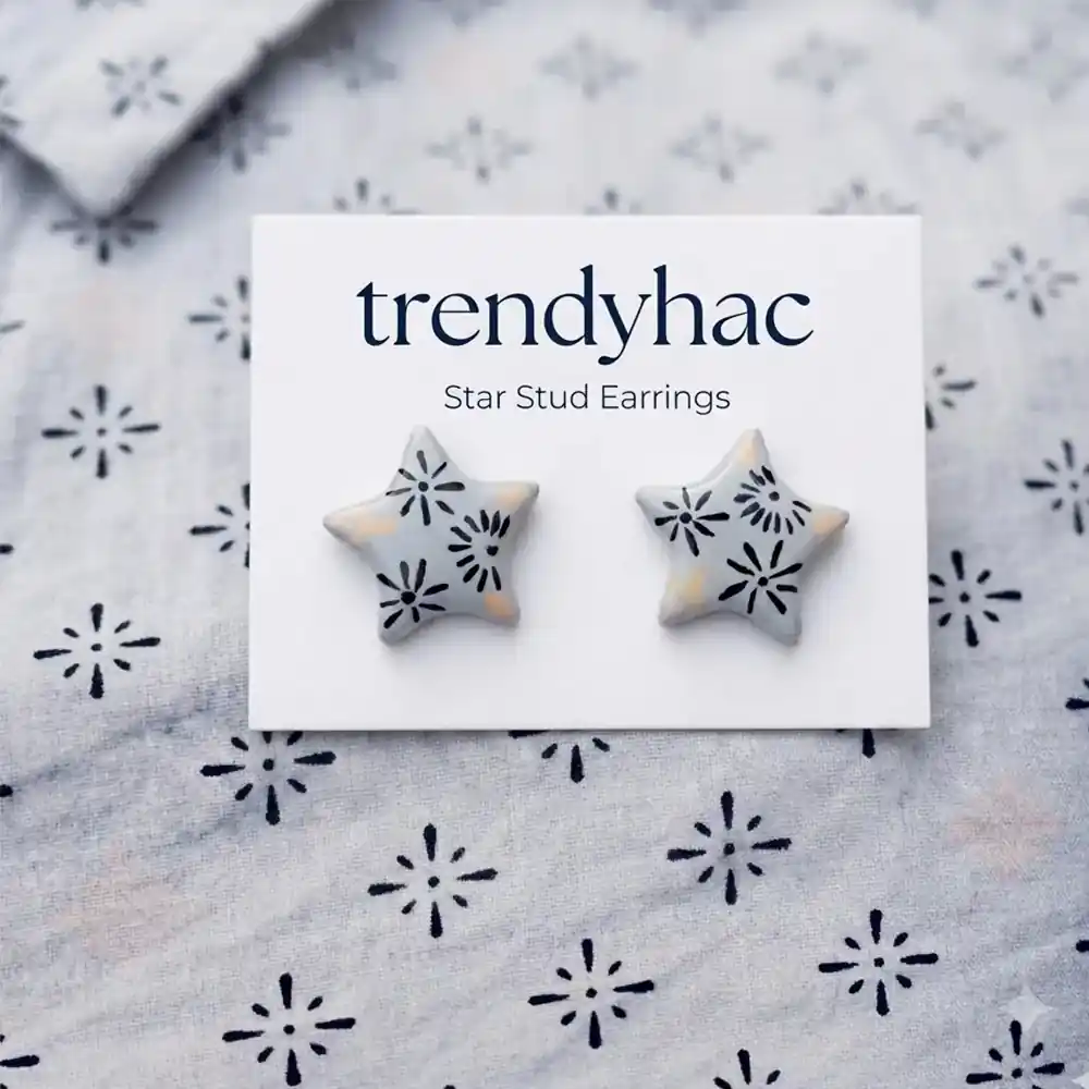 Flat lay view of light grey star-shaped clay stud earrings with hand-painted black starburst patterns on a white TRENDYHAC display card matching the fabric background.