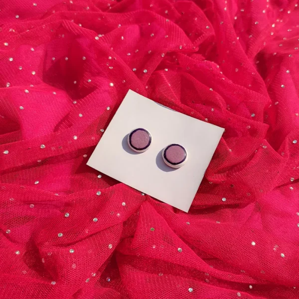 Flat lay view of a pair of circular purple stud earrings on a white display card set against bright pink glittery mesh fabric.