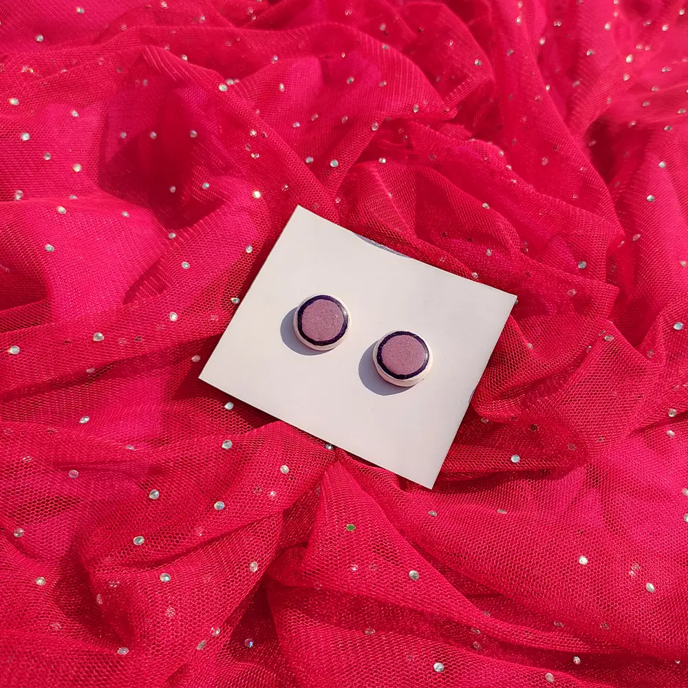 Flat lay view of a pair of circular purple stud earrings on a white display card set against bright pink glittery mesh fabric.