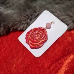 Flat lay view of a red sculpted polymer clay rose pendant with gold highlights and a pink bead on a white card over red and grey fur fabric.