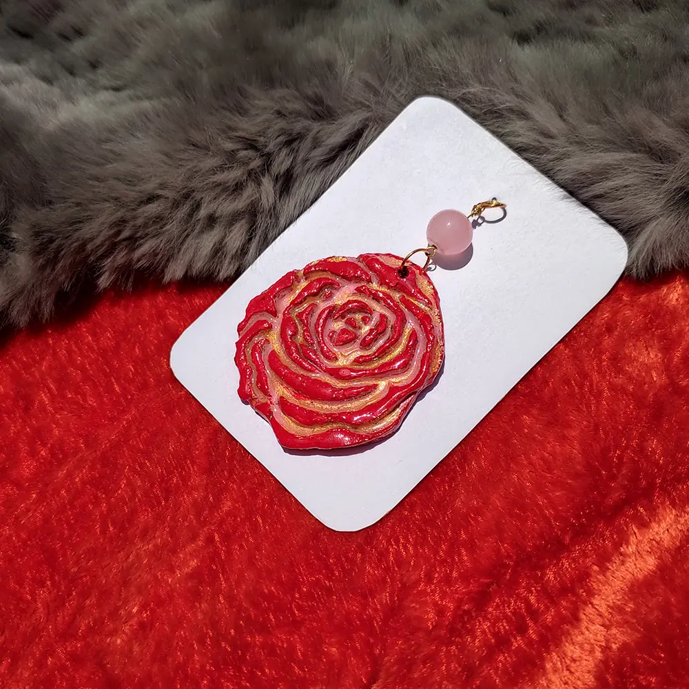 Flat lay view of a red sculpted polymer clay rose pendant with gold highlights and a pink bead on a white card over red and grey fur fabric.