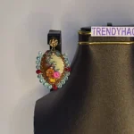 Close-up of an oval clay earring featuring 3D sculpted orange and pink flowers with green vines, bordered by sky blue and red crystal beads on a black bust.