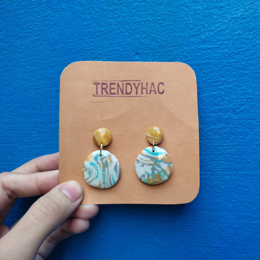 Handheld view of a tan Trendyhac display card holding a pair of circular marbled drop earrings with teal, white, and gold swirls against a blue wall.