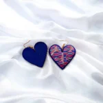 Flat lay view showing the decorated front face of one heart earring next to the solid, textured dark blue reverse side of the matching piece on white fabric.