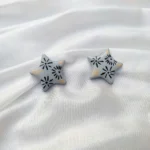 Flat lay view of a pair of grey five-point star stud earrings with black floral patterns on a white satin fabric background.