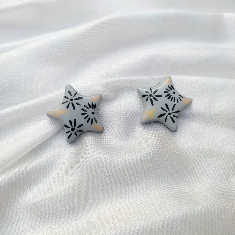 Flat lay view of a pair of grey five-point star stud earrings with black floral patterns on a white satin fabric background.