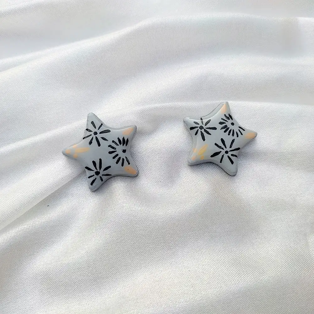 Flat lay view of a pair of grey five-point star stud earrings with black floral patterns on a white satin fabric background.