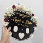 Close up view of a round black acrylic thumb impression board with gold lettering and a crescent border of artificial flowers.