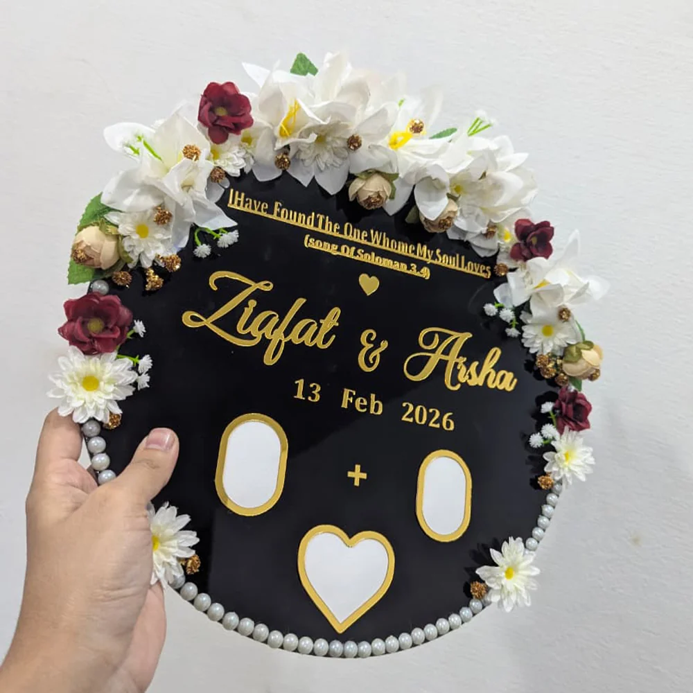 Close up view of a round black acrylic thumb impression board with gold lettering and a crescent border of artificial flowers.