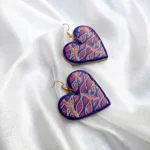 A pair of glossy heart-shaped clay earrings with a pink and blue wave pattern and crane illustrations, resting on soft draped white silk fabric.
