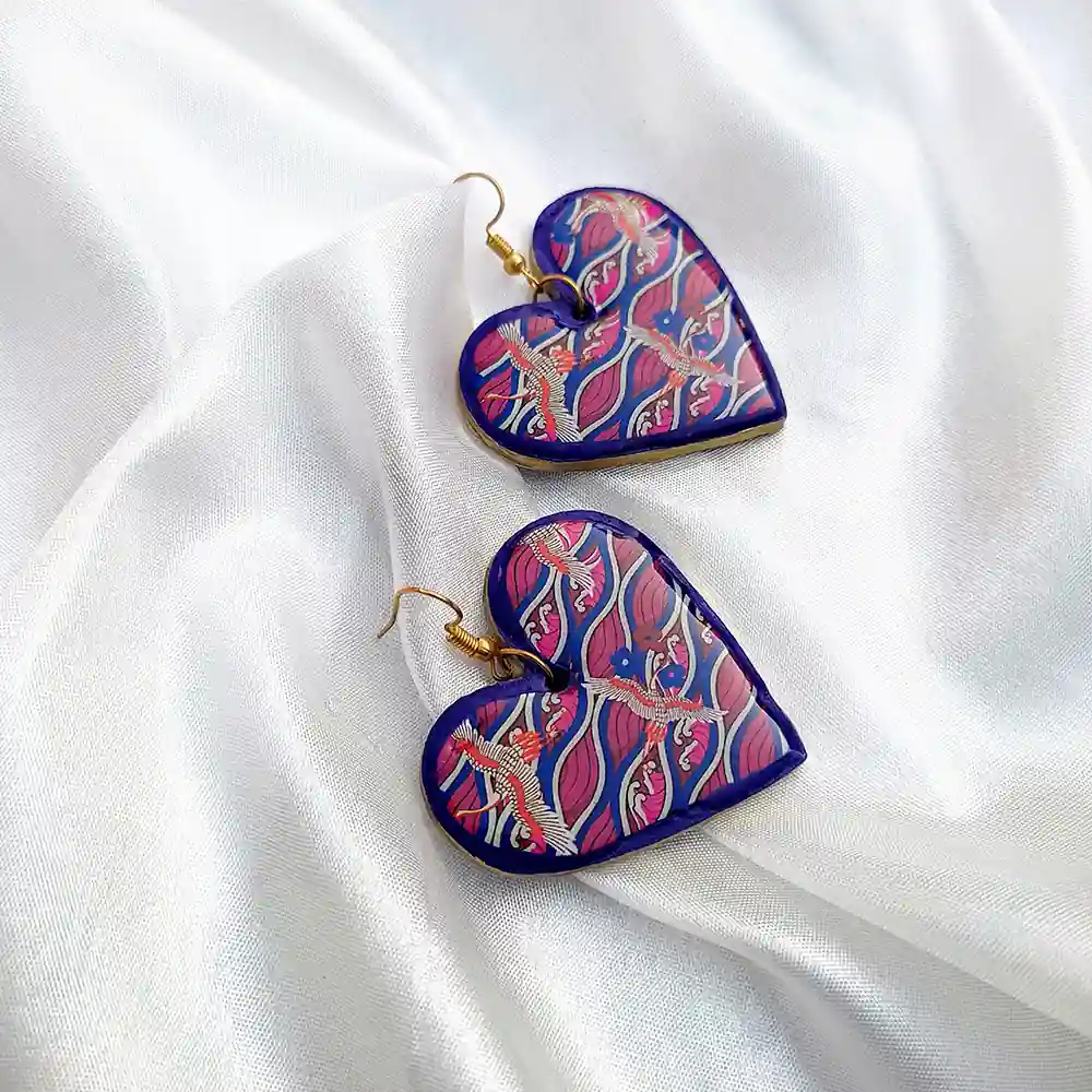 A pair of glossy heart-shaped clay earrings with a pink and blue wave pattern and crane illustrations, resting on soft draped white silk fabric.