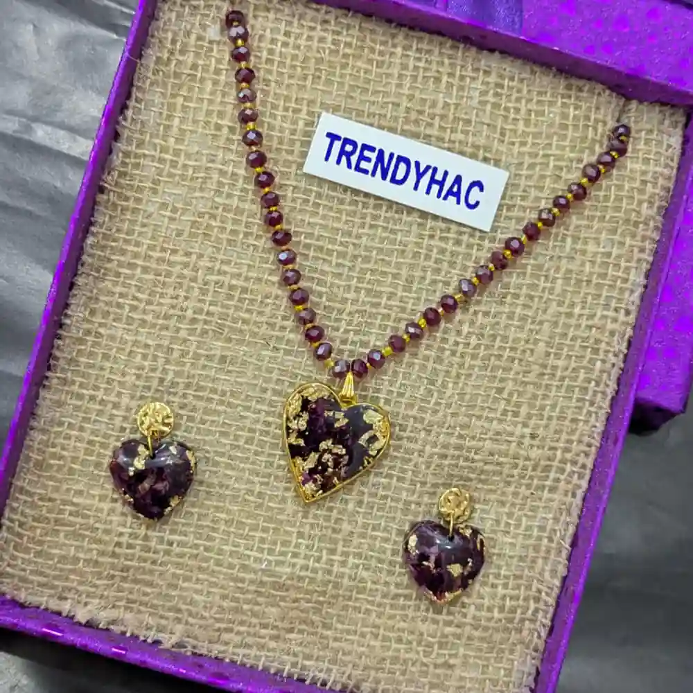 Preserved red rose petal heart pendant on a beaded necklace beside matching drop earrings in a purple box.